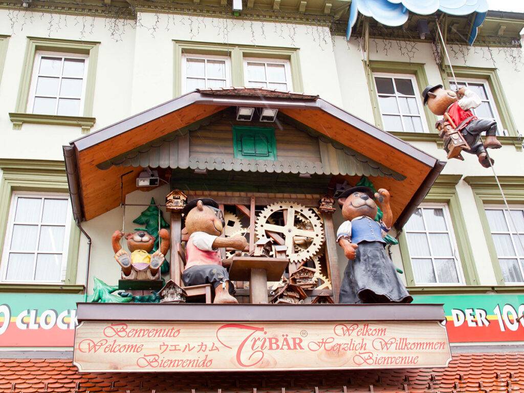 An oversized cuckoo clock on the front of a building. Several dressed teddy bears sit in front of it.