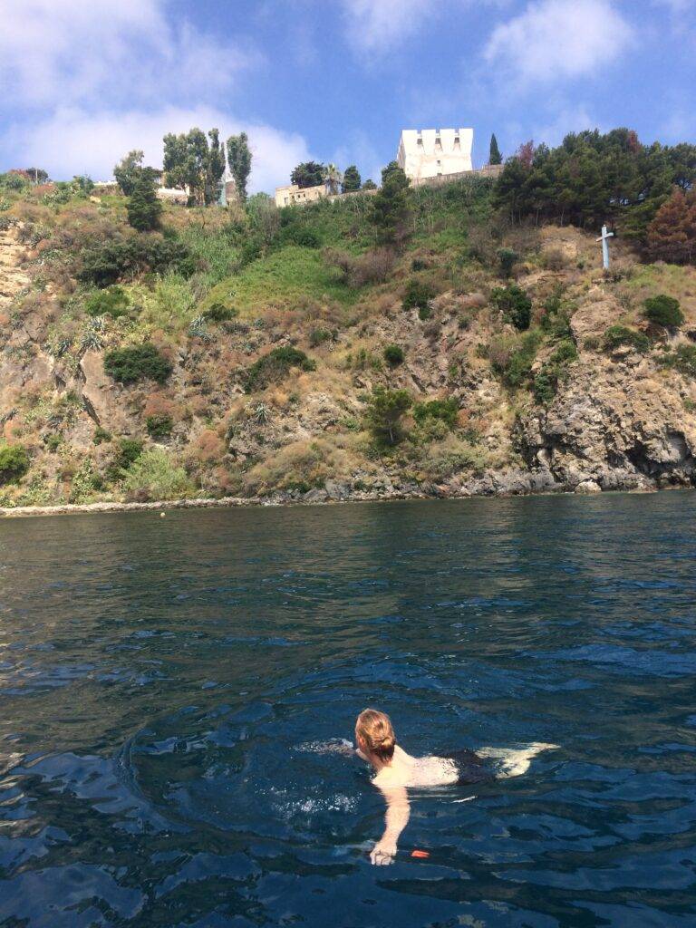A man with red hair swimming in the sea, looking up at a building on the top of a grassy cliff