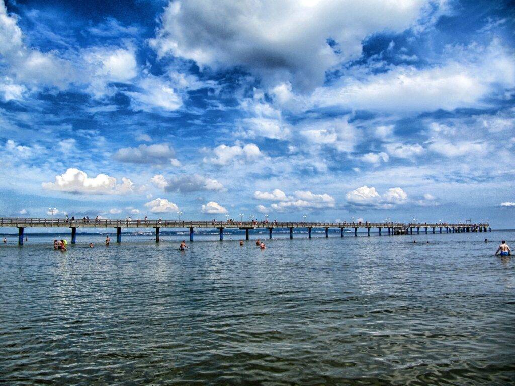 A long pier stretching out to the sea with a blue cloudy sky in the background and swimmers in the water